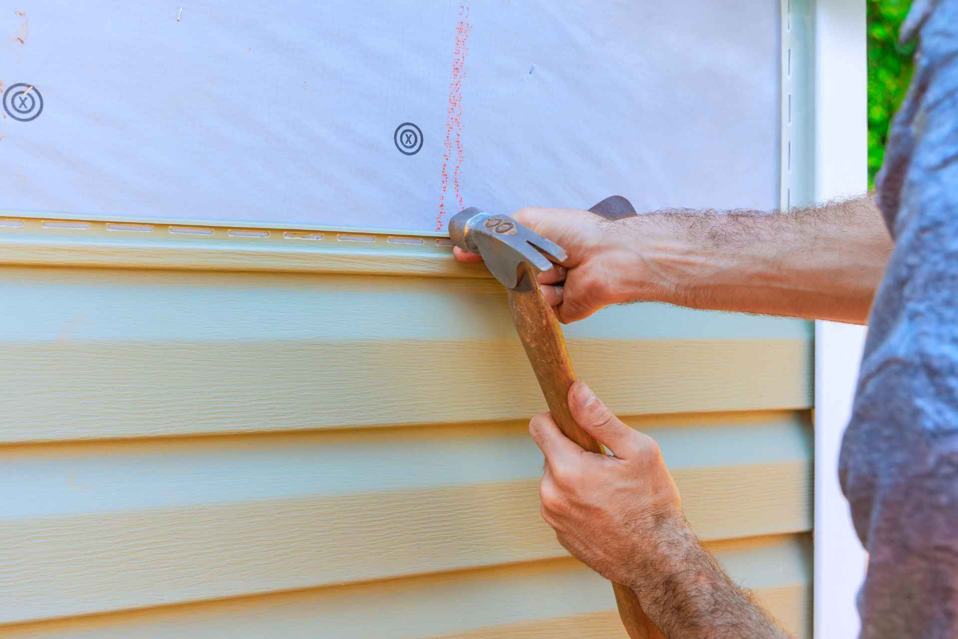 Close-up of a construction worker's hands holding a hammer to carefully install new exterior siding along a residential wall during a fall renovation.